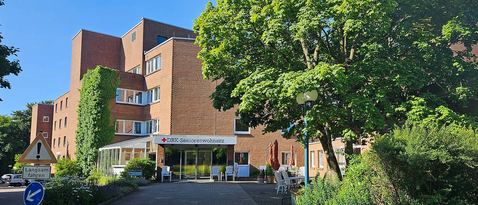 Blick auf ein großes rotes Backsteingebäude. Über dem Eingang steht auf einem weißen Schild: DRK-Seniorenwohnsitz. Rechts vor dem Eingang sowie links neben dem Gebäude stehen Bäume.
